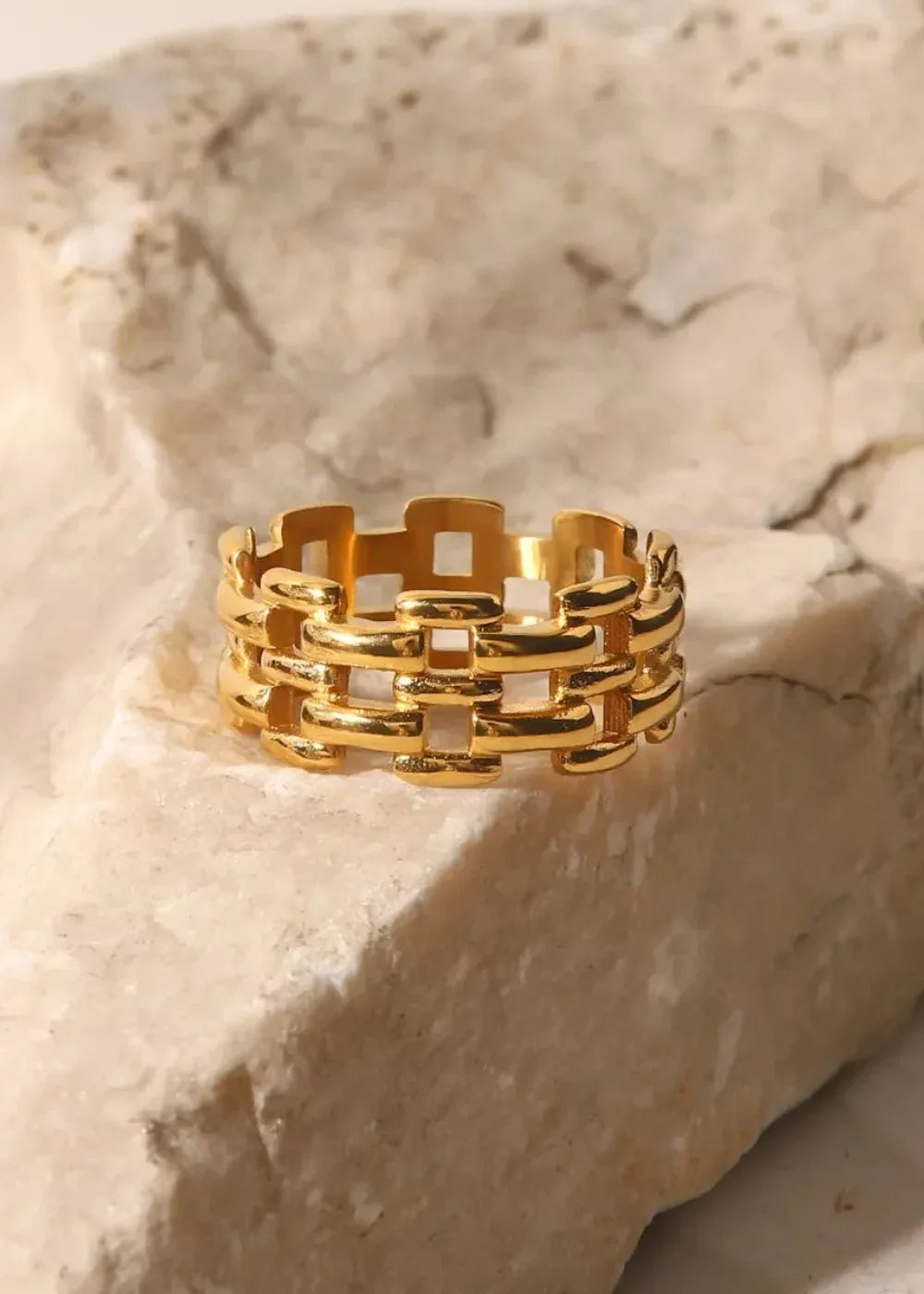 Gold plated stainless steel ring with a woven link pattern rests on a textured stone surface.