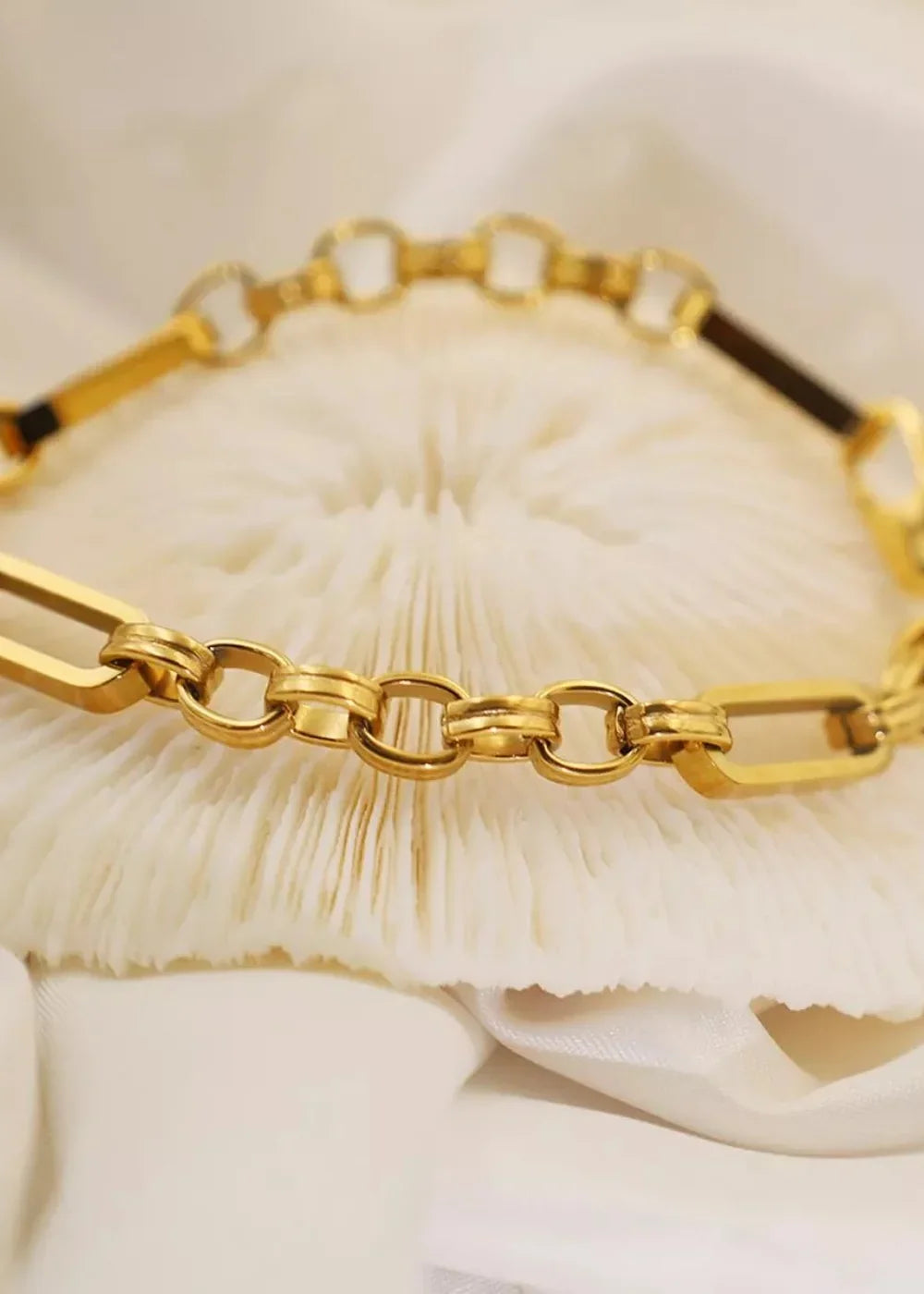 Close-up of a gold chain bracelet with interlocking oval and circular links resting on a textured white surface.