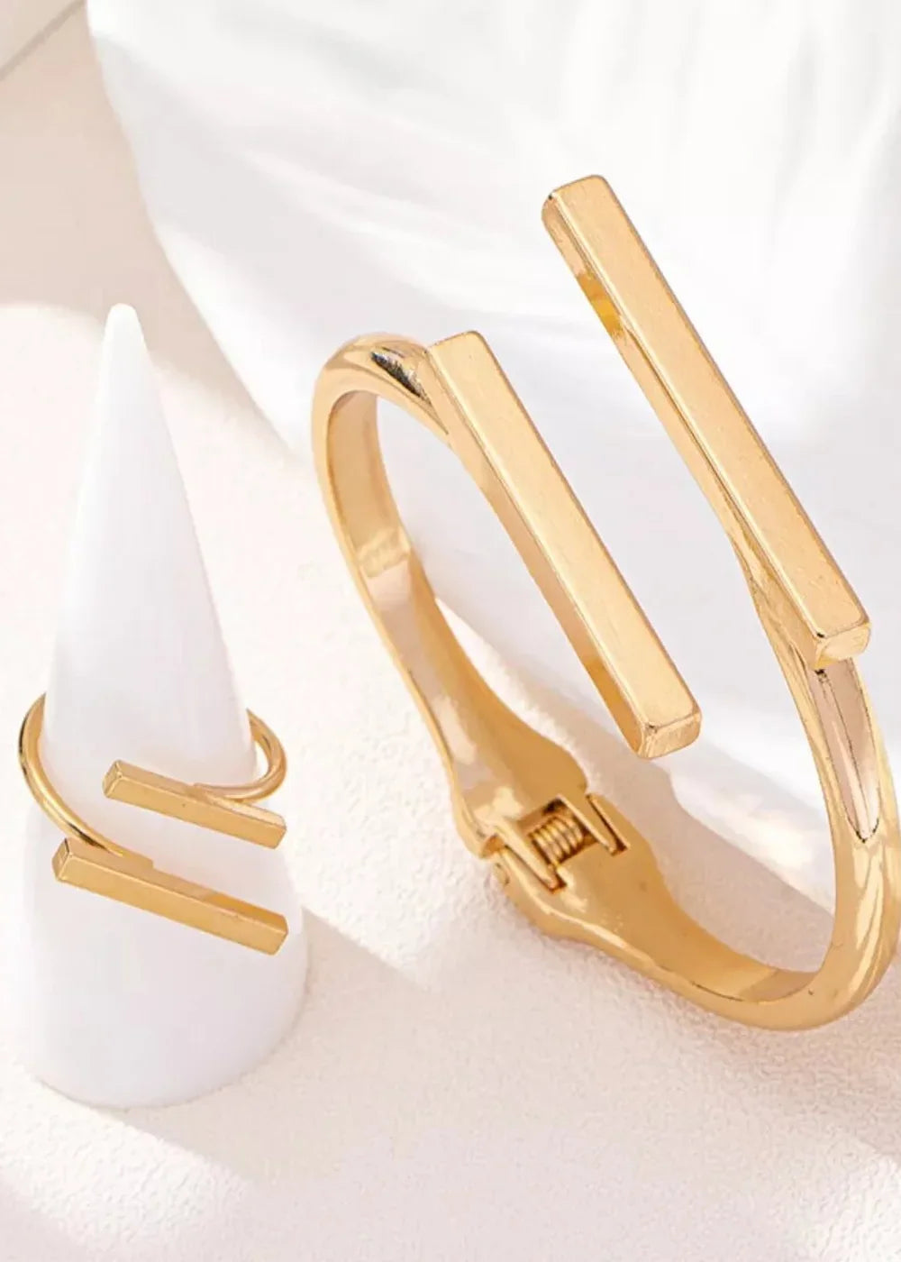 Gold-plated open cuff bracelet and minimalist bar ring on a white cone display.