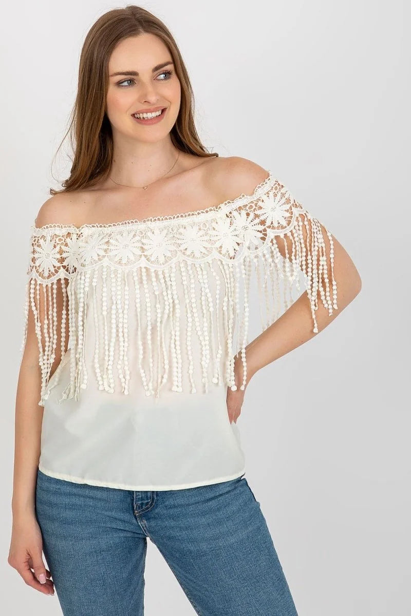 Off-the-shoulder ivory lace top with fringe detail and white beaded accents, worn with blue jeans.