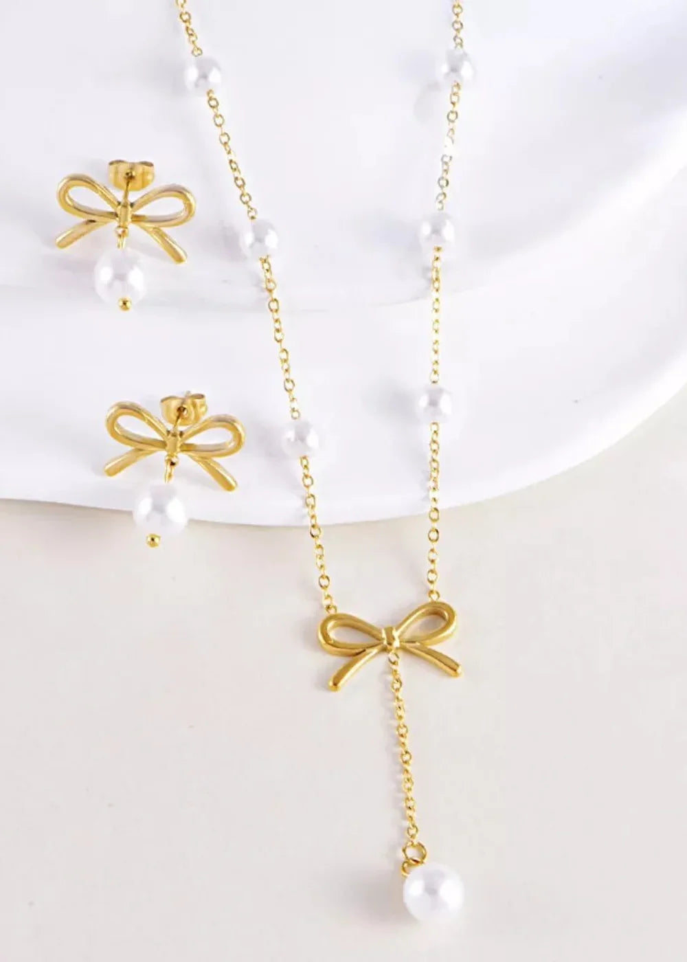 Gold bow necklace and earrings with pearls, on a white surface.