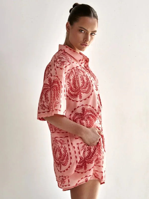 Pink short suit with a red palm tree print, worn by a model.