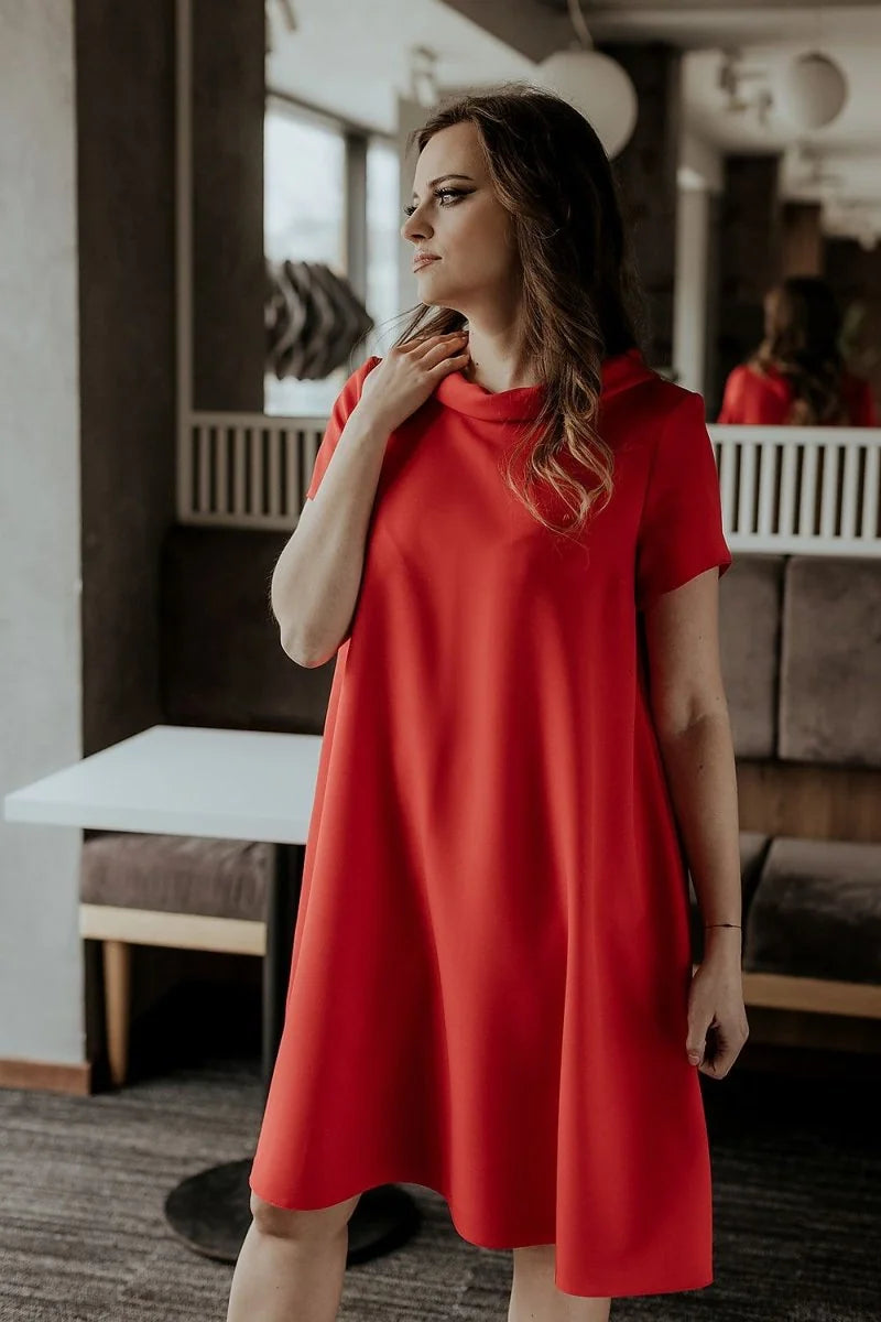 A woman in a red loose-fit day dress with short sleeves stands looking to the side.