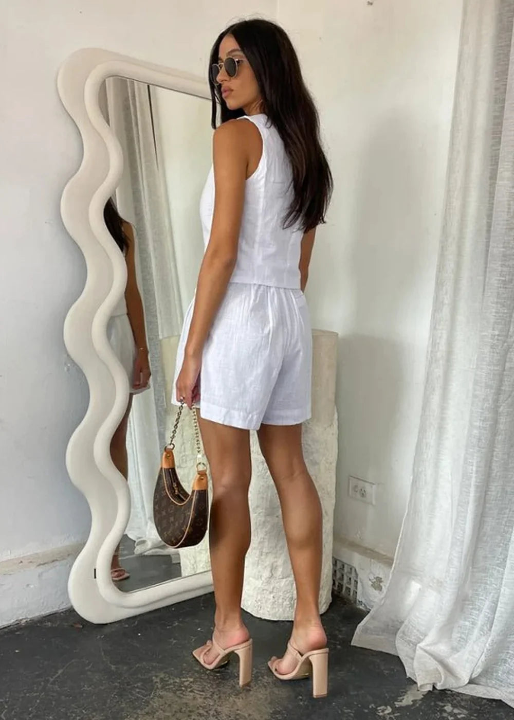 Woman in a white linen sleeveless top and shorts, holding a brown monogrammed shoulder bag and wearing nude heeled mules.