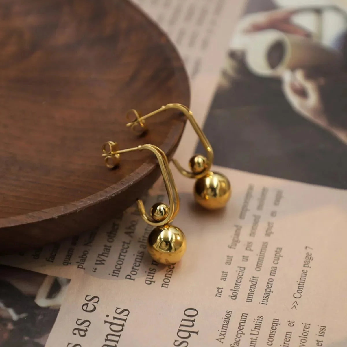 Close-up of shiny gold earrings with spherical charms, resting on a wooden surface and newspaper.