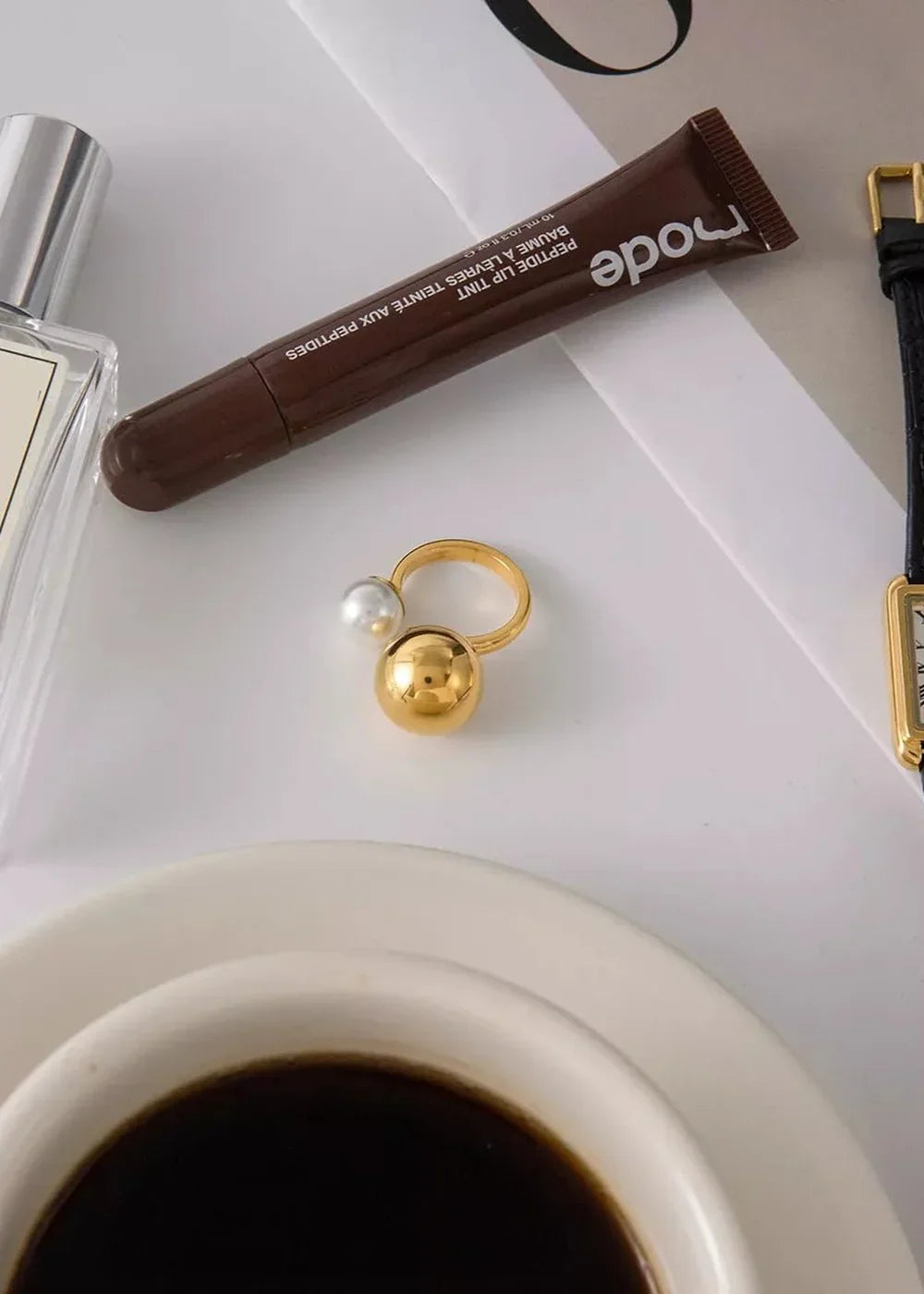 Gold ring with large pearl and a gold sphere, next to a brown lip tint tube.