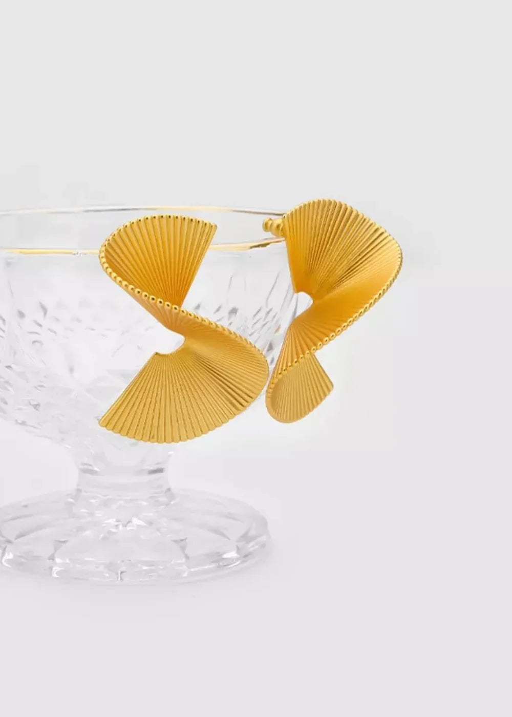 Shiny gold-plated fan-shaped earrings with ribbed texture against a clear glass bowl.