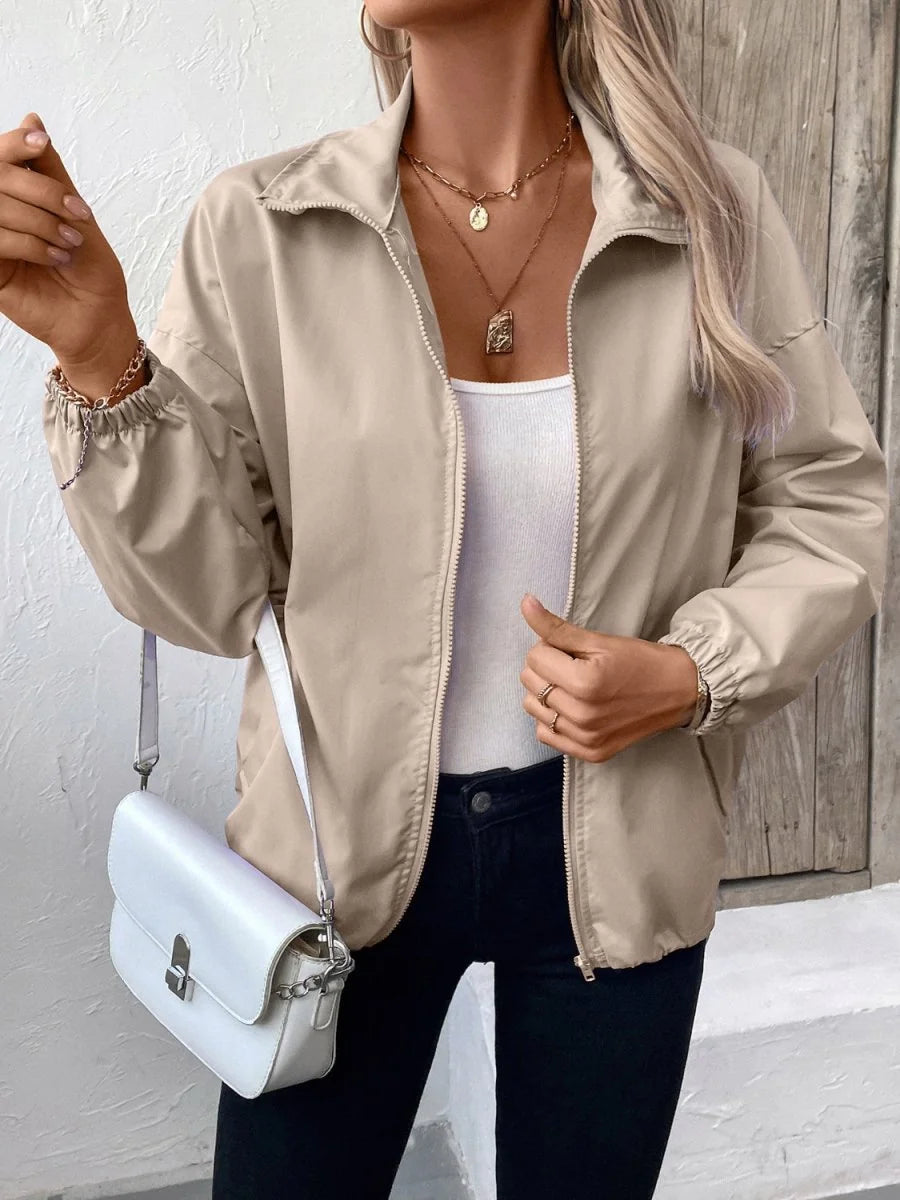 A woman wears a beige zippered jacket with a white tank top, black jeans, and a white purse.