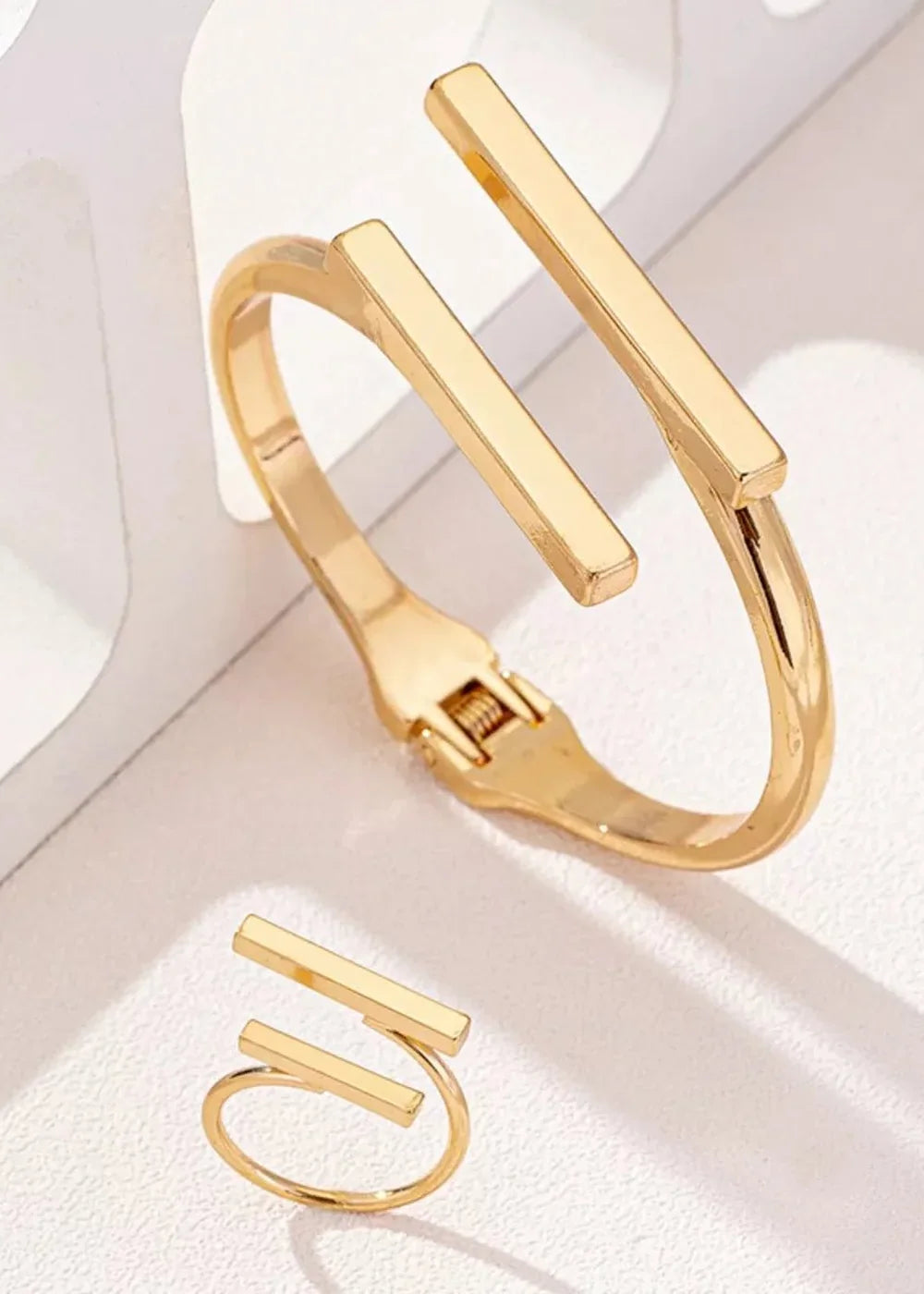 Gold-plated cuff bracelet with linear bar accents and matching ring, displayed on a white surface.