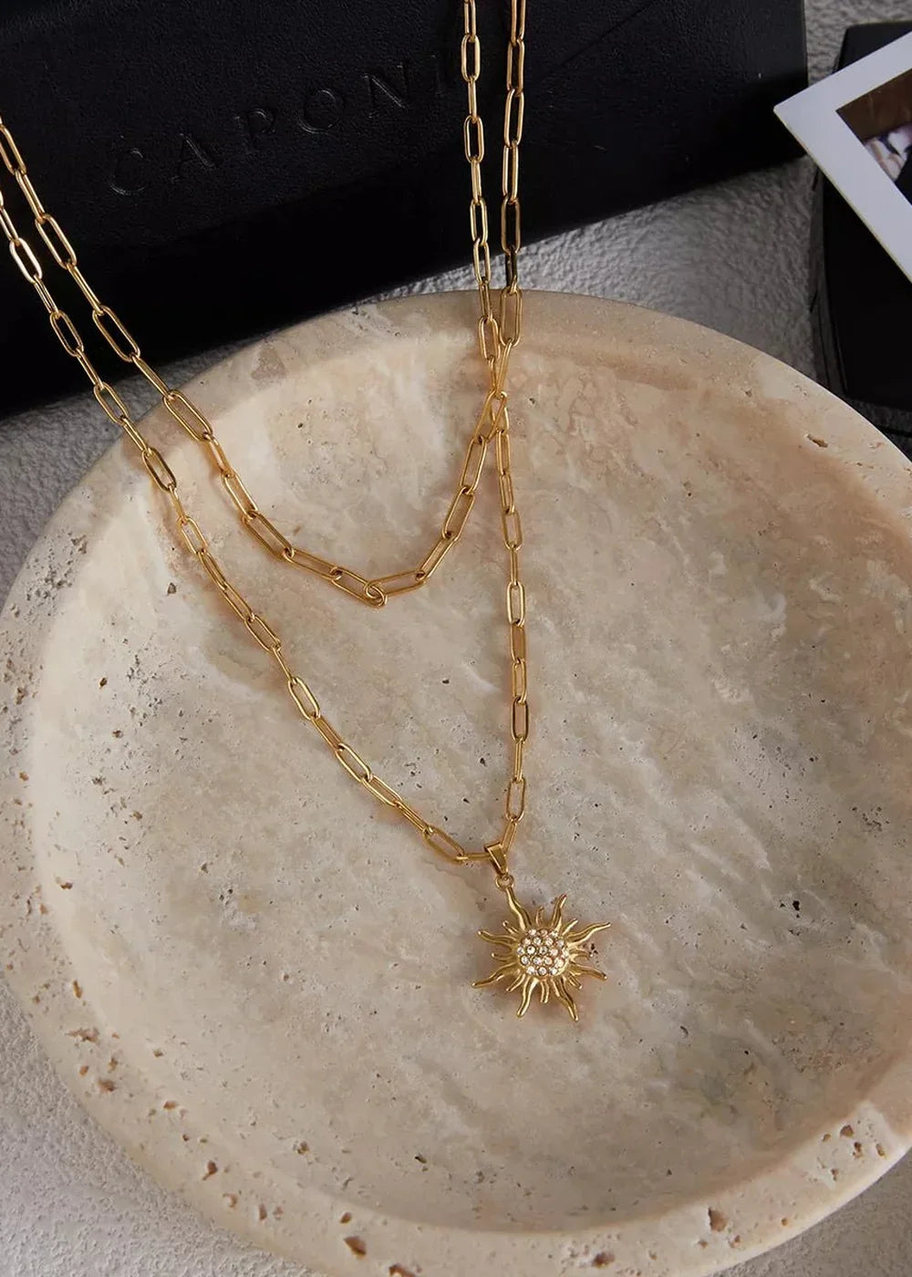 Gold layered necklace with a sparkling sun pendant resting on a textured stone dish.