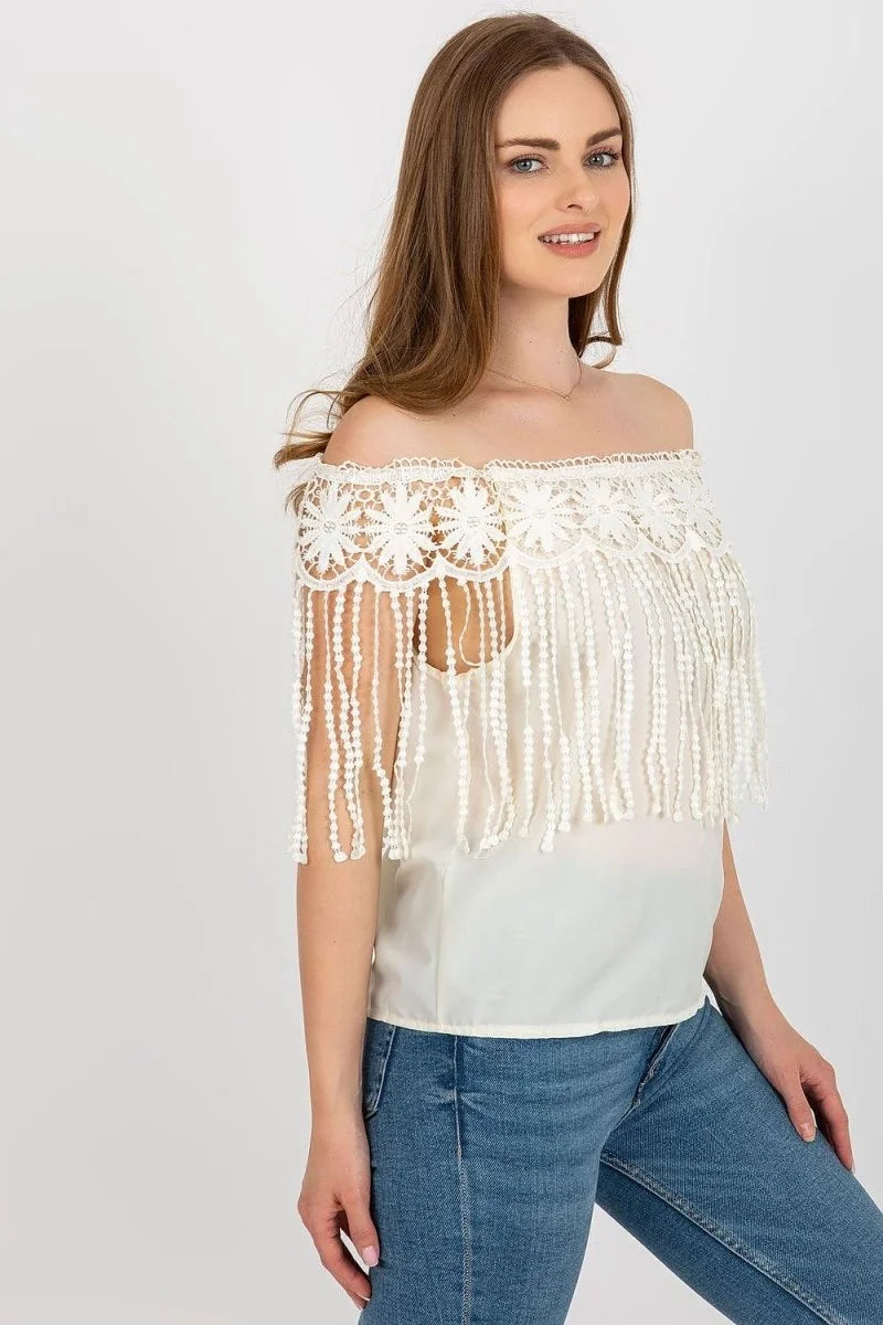 Off-white crochet off-the-shoulder top with fringe detail over a camisole, paired with blue jeans.