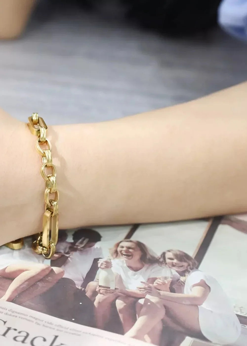 Close-up of a gold-plated chain bracelet on a wrist, with a magazine spread of laughing women in white t-shirts below.