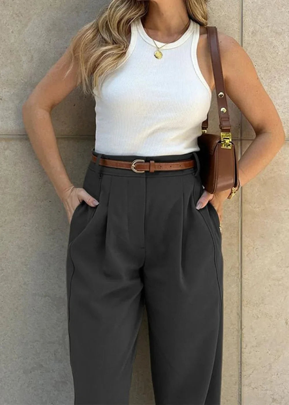 Woman wearing dark gray pleated pants and a white ribbed tank top, accessorized with a brown belt and brown shoulder bag.