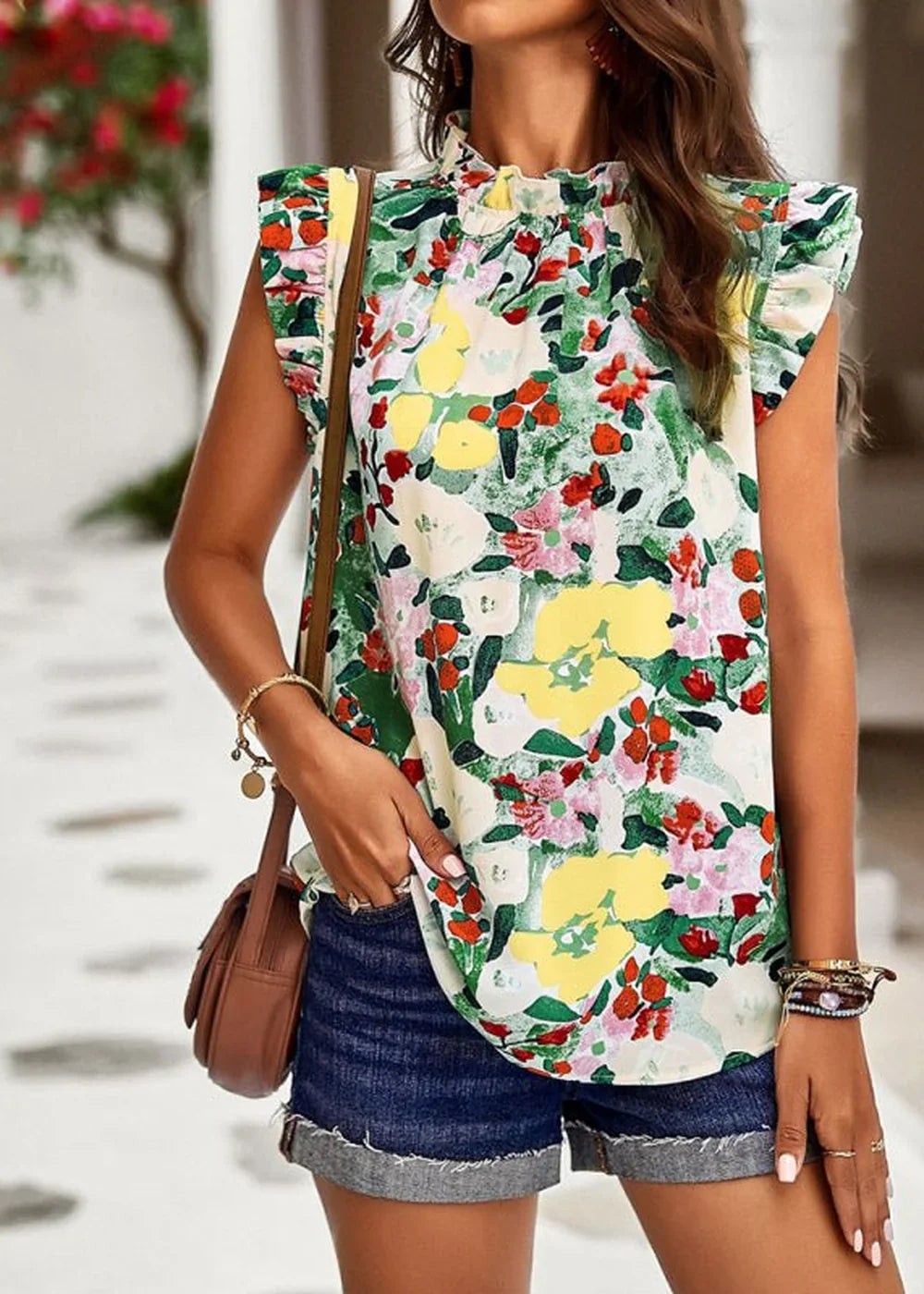 Woman in a floral print ruffled mock neck top and denim shorts, holding a brown crossbody bag.