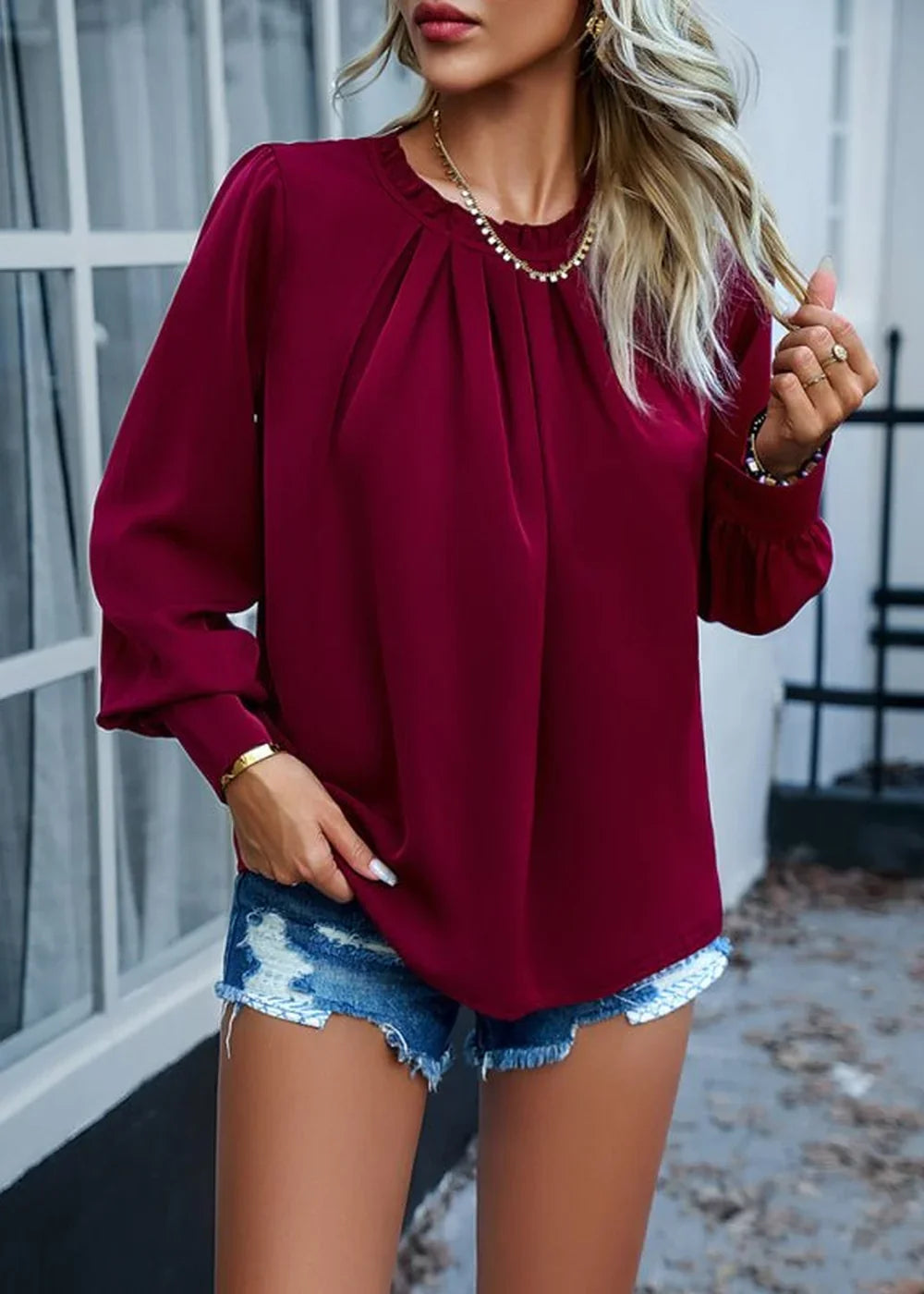 Burgundy pleated long-sleeve top with gold jewelry and distressed denim shorts on model