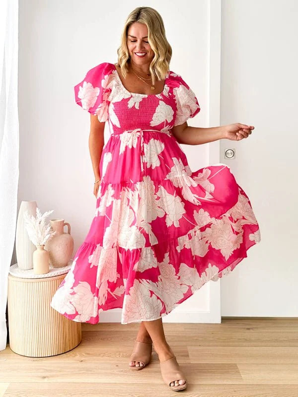 Vibrant pink tiered midi dress with a white floral print, puff sleeves, and a smocked bodice on a smiling model.