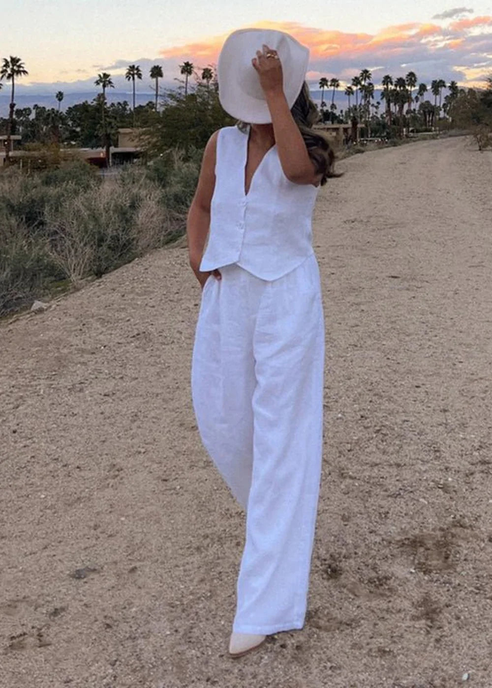 Woman in white linen vest and pants, wearing a white hat, standing outdoors at sunset.