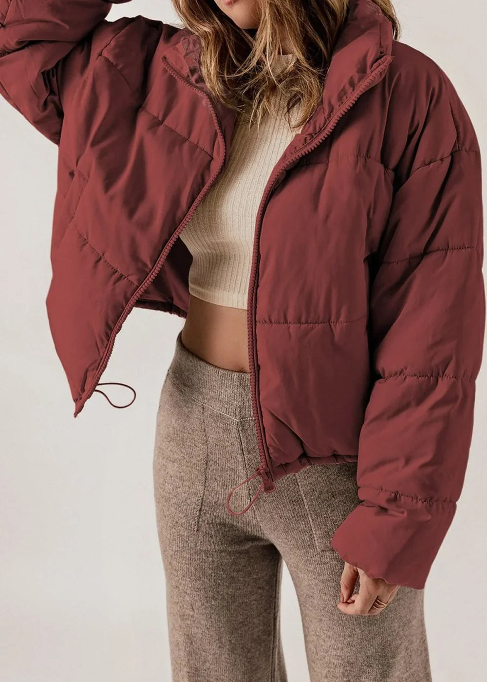 A maroon puffer jacket with a high collar and zipper closure, worn over a cream ribbed crop top and brown knit pants.