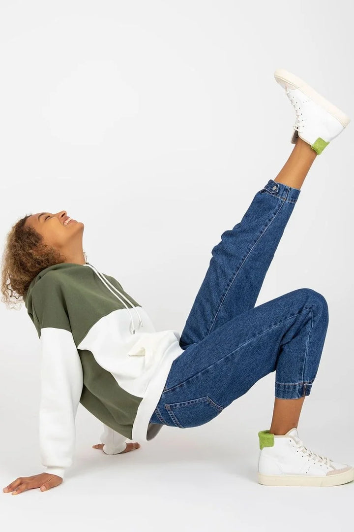 A woman in a colorblock hoodie and blue jeans is in a playful pose, with one leg extended upwards.