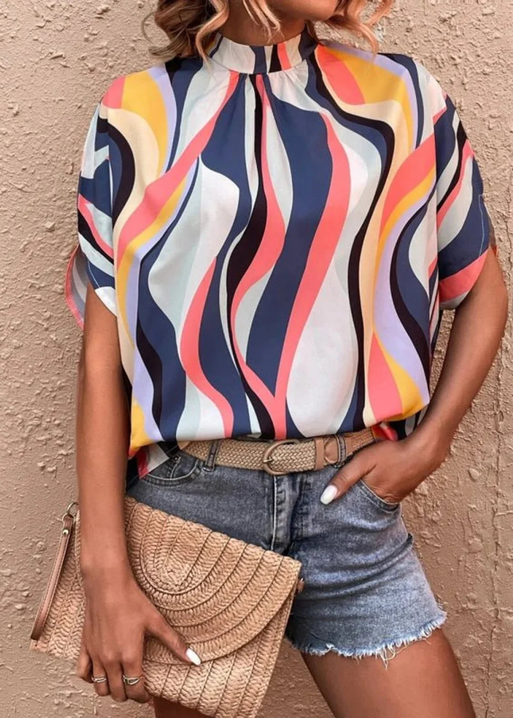 A woman wears a short-sleeved, high-neck blouse with a colorful wavy print, paired with distressed denim shorts and a wove...