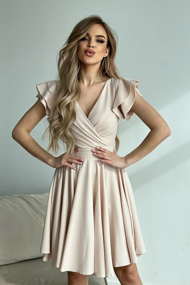 A woman in a light peach wrap-style cocktail dress with ruffled sleeves poses with hands on hips.