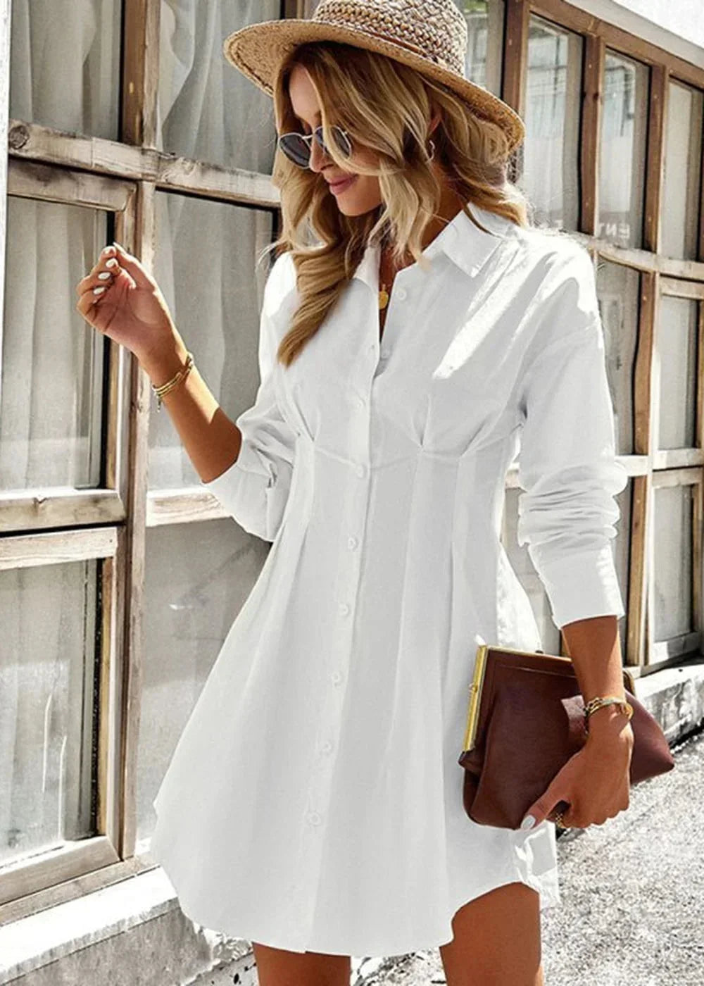 Woman in a white button-down shirtdress with a cinched waist and a straw hat, holding a brown clutch.