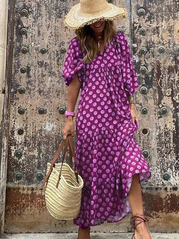 Purple polka dot maxi dress with a V-neck, tiered skirt, and straw hat, held by a straw bag.