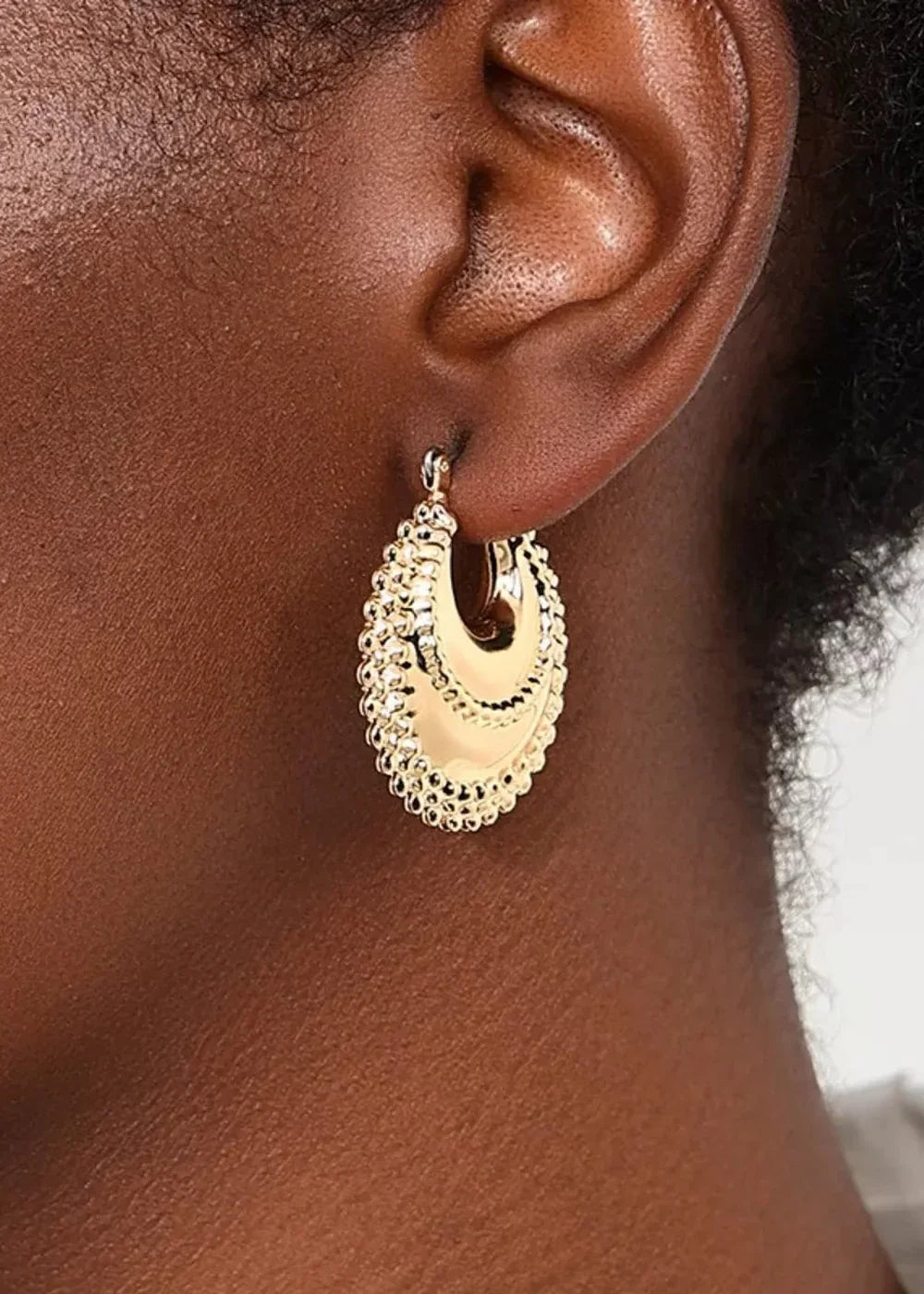 Close-up of a gold hoop earring with a textured border on a dark skin tone.
