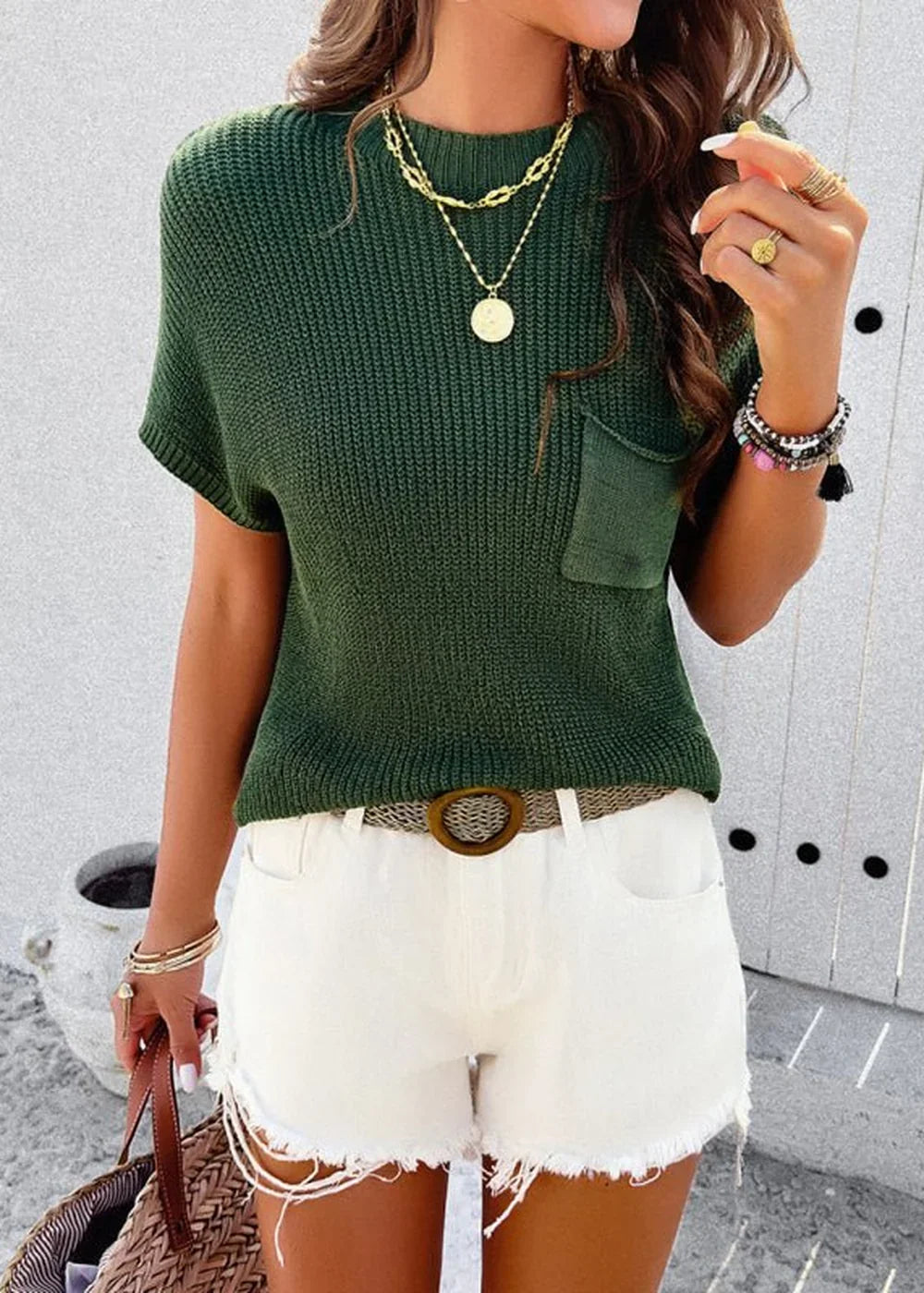 Woman wearing a dark green knit top with a pocket, white distressed shorts, and a woven belt.
