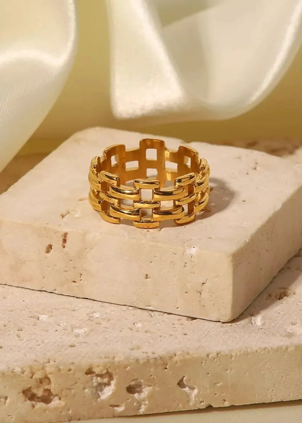 Gold-toned stainless steel ring with a woven, interlocking band design, resting on a textured stone surface.