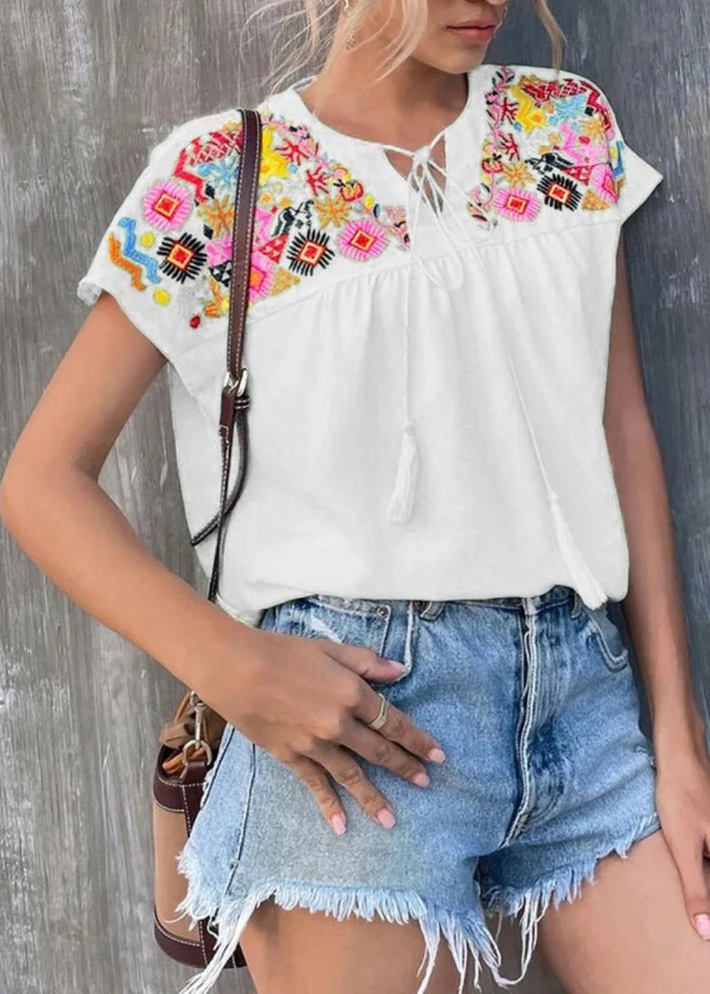 White embroidered top with colorful floral accents and tie detail, paired with distressed denim shorts.