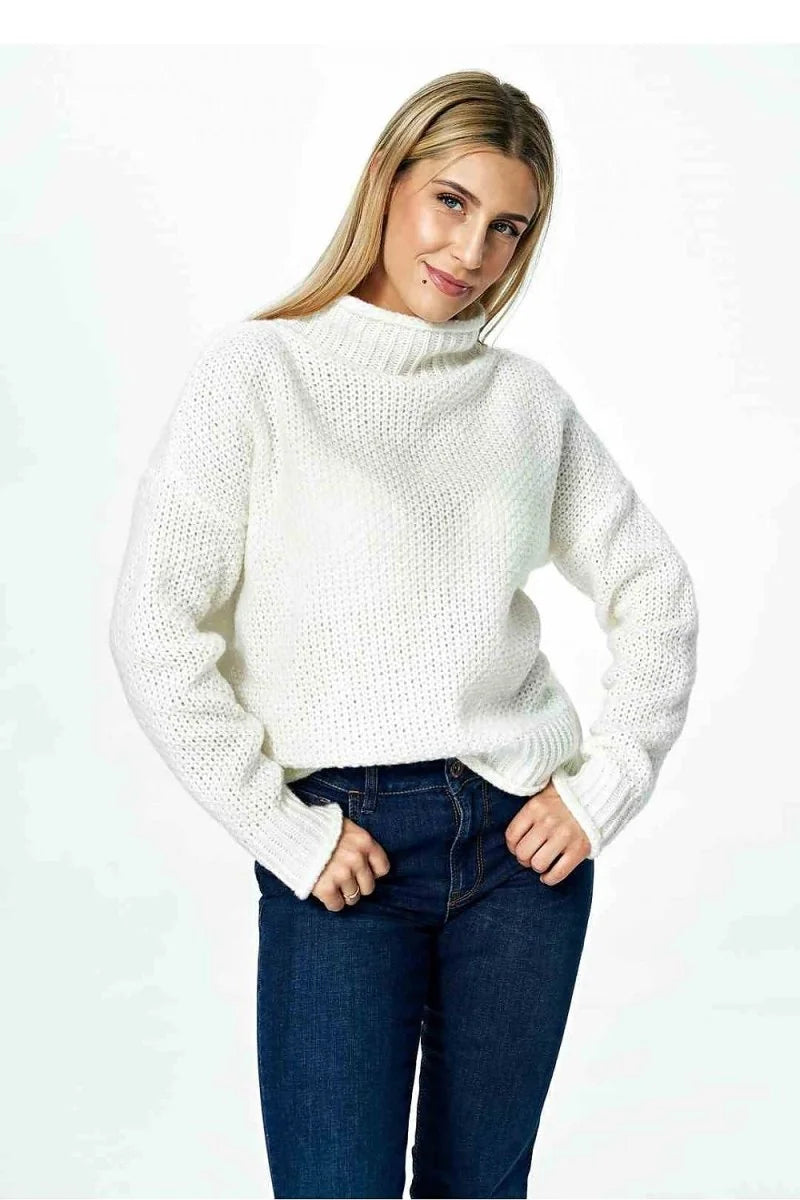 A woman in a white knit turtleneck sweater and dark wash jeans, hands in pockets.