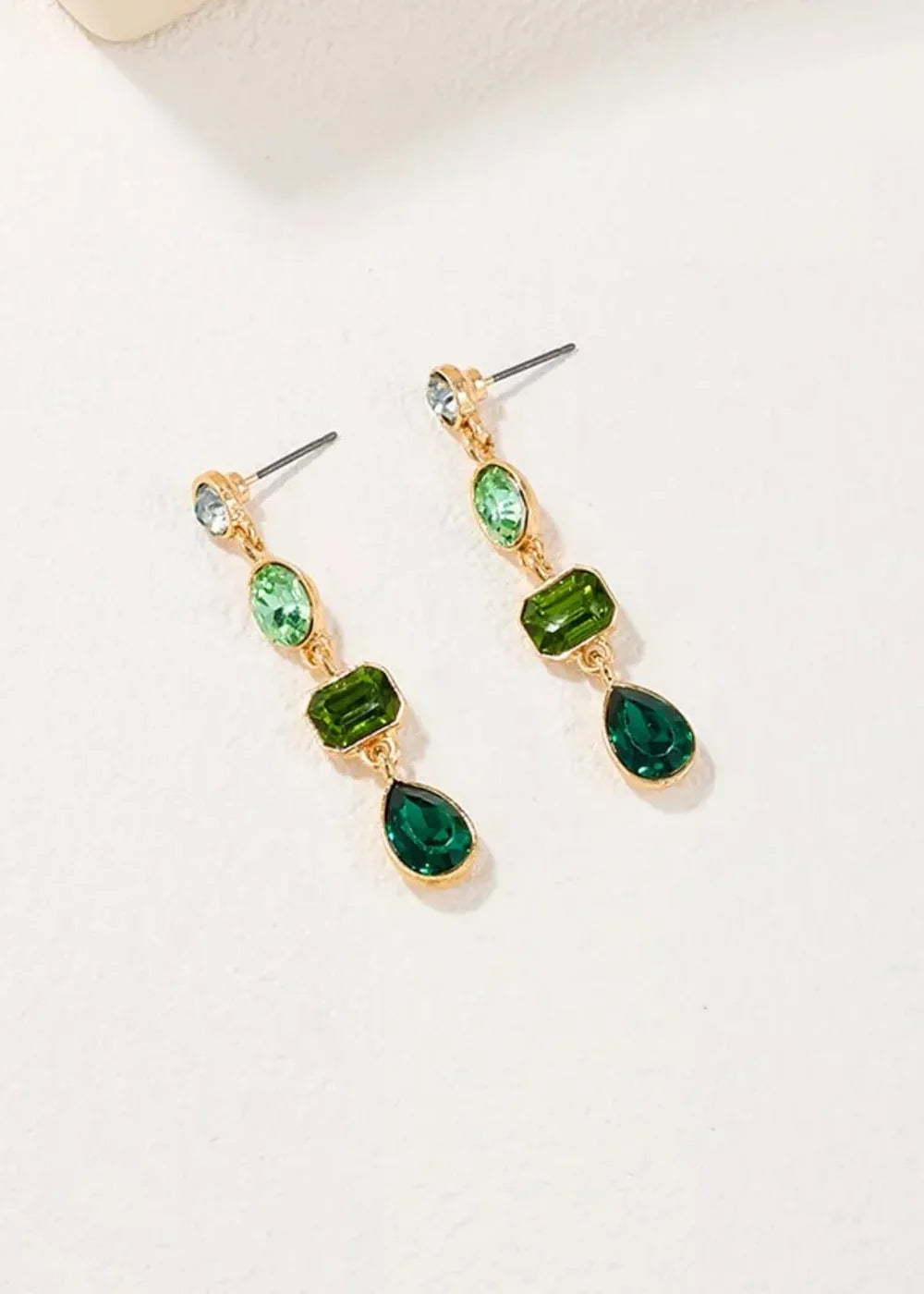Gold-toned dangle earrings with clear, light green, and emerald green faceted stones in oval, rectangular, and teardrop sh...