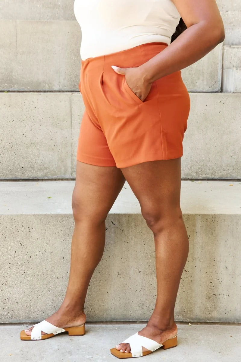 Caramel orange pleated shorts with pockets, worn by a model with white slide sandals.
