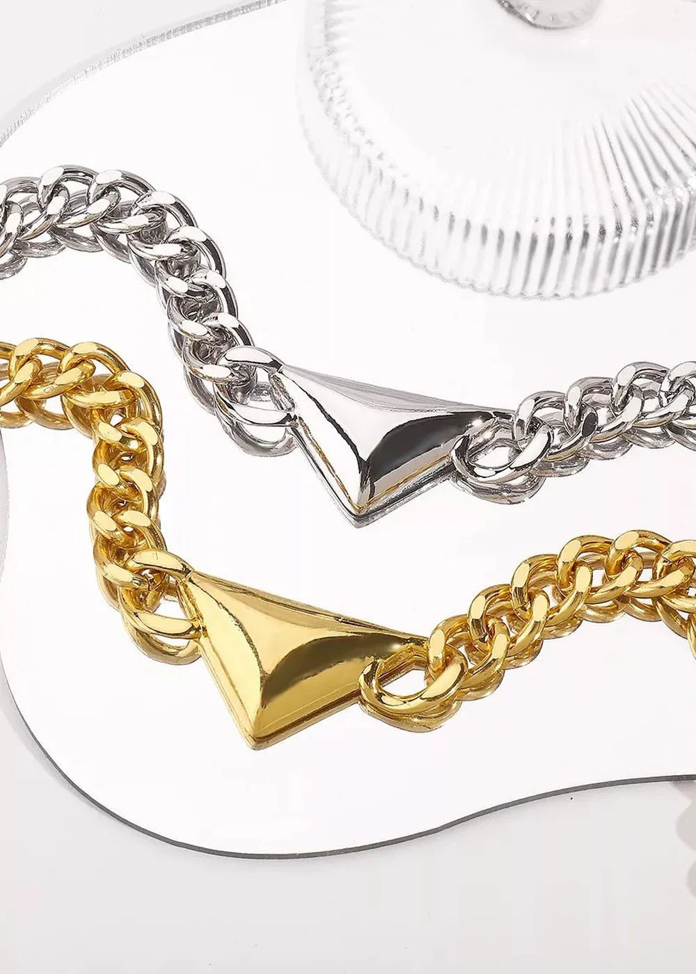 Gold and silver chunky chain bracelets with triangular charms, reflected on a mirrored surface.