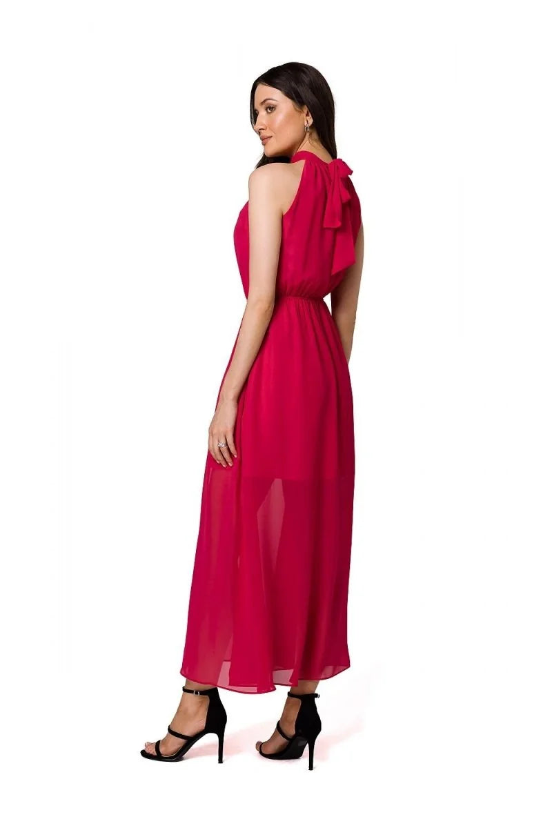 Model in a fuchsia halter neck maxi dress with a bow detail at the neck and sheer skirt, wearing black strappy heels.