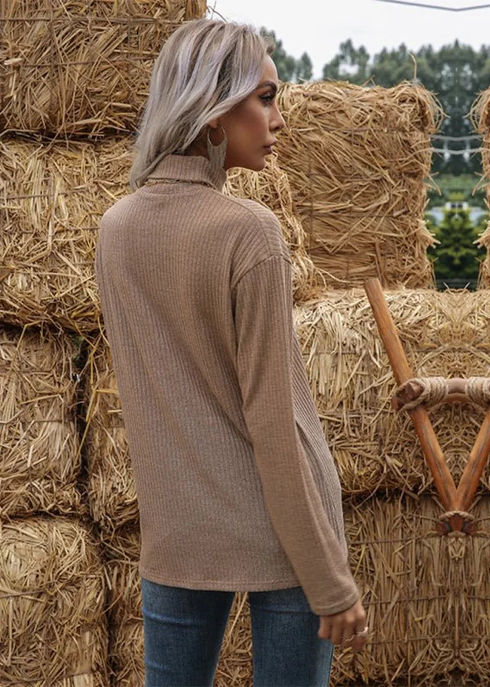 Woman in a tan ribbed turtleneck sweater with blue jeans, standing in front of hay bales.