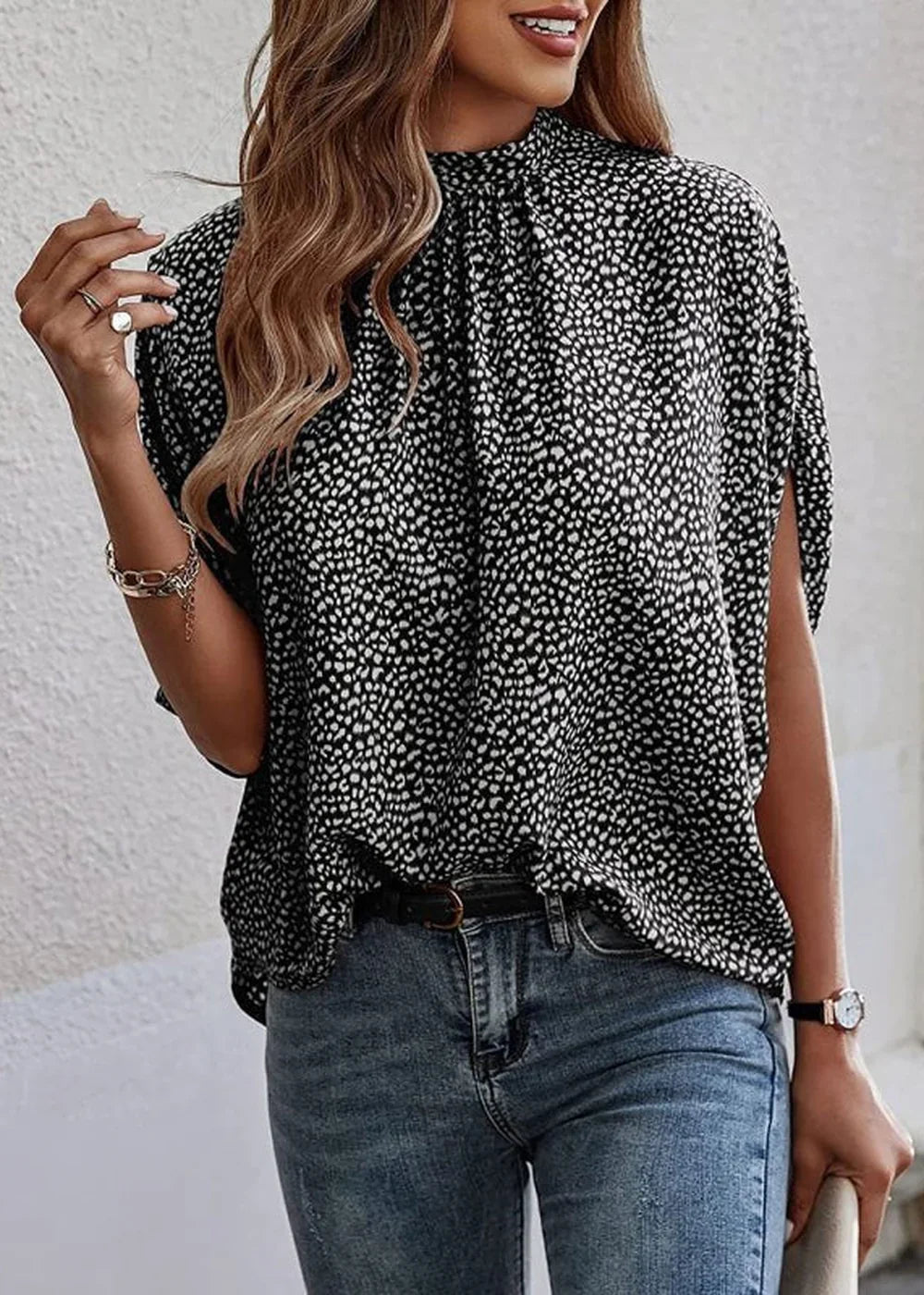 Black and white speckled print mock neck top with split sleeves, worn with blue jeans and a belt.