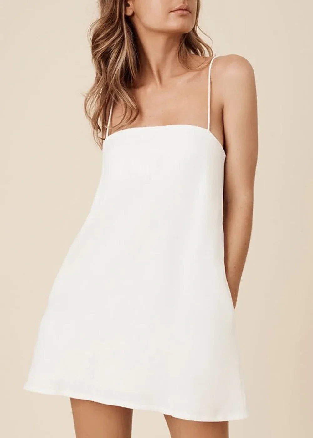 Model in a white cotton and linen blend slip dress with thin spaghetti straps and a square neckline.