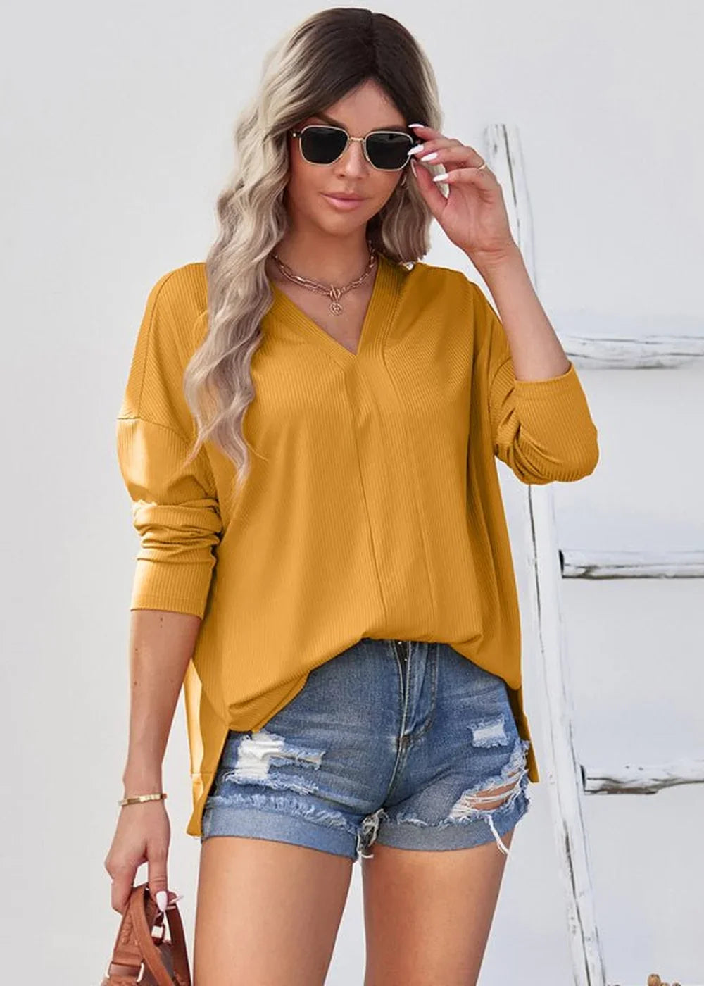 Woman in mustard yellow ribbed v-neck top and distressed denim shorts, wearing sunglasses and necklaces.