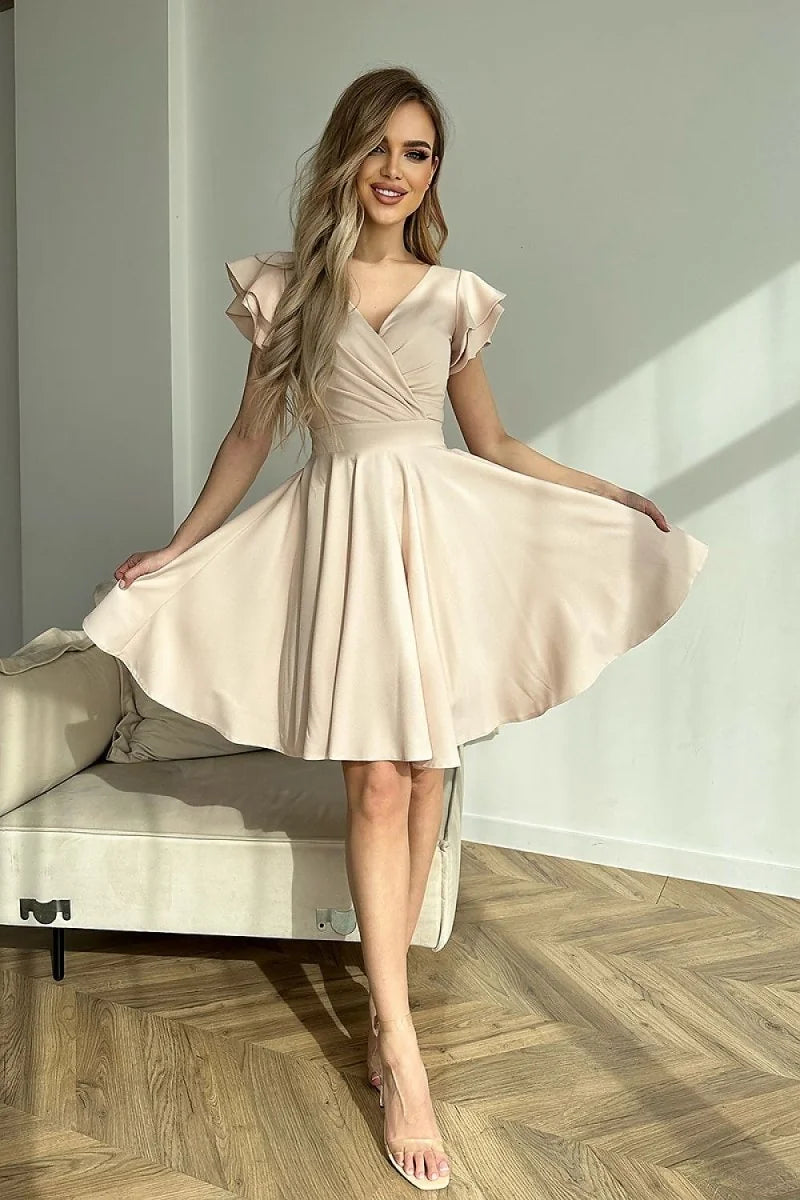 A woman in a flowing, champagne-colored cocktail dress with ruffled short sleeves and a V-neck, standing on a wooden floor.