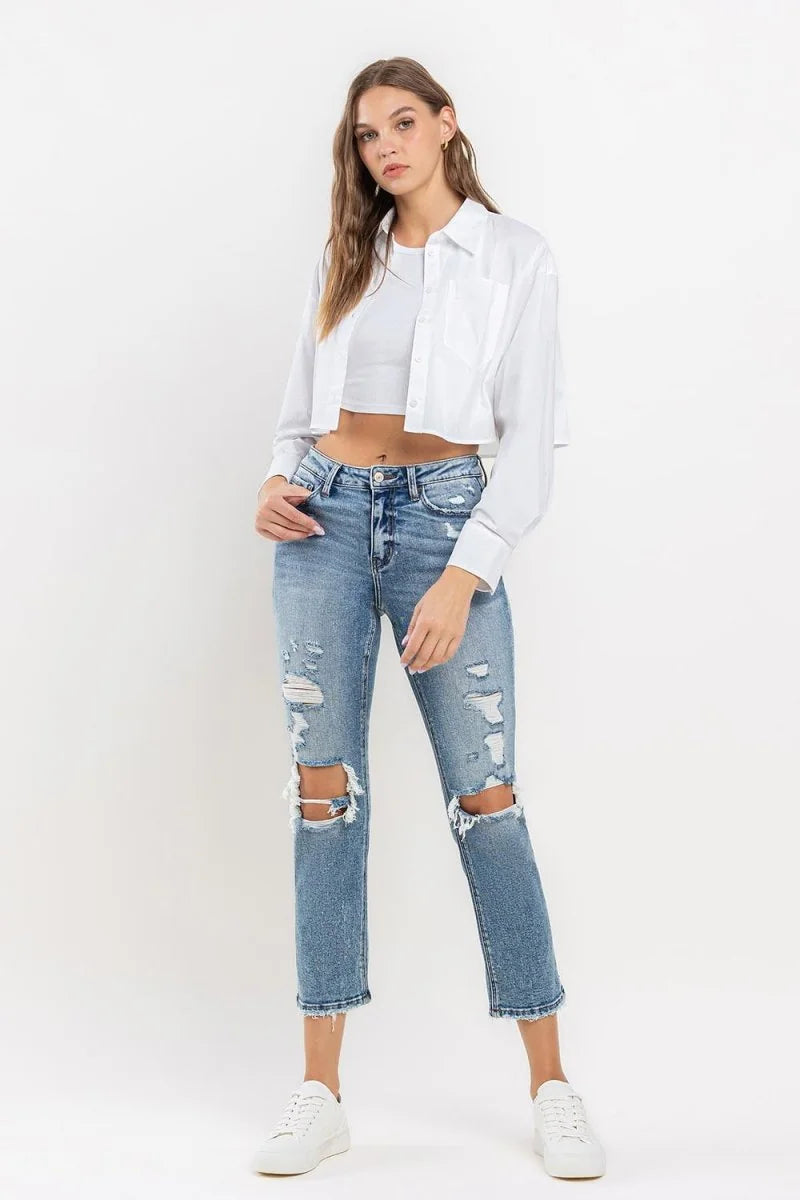 Distressed medium-wash denim jeans with ripped knees and frayed hem, worn by a model.