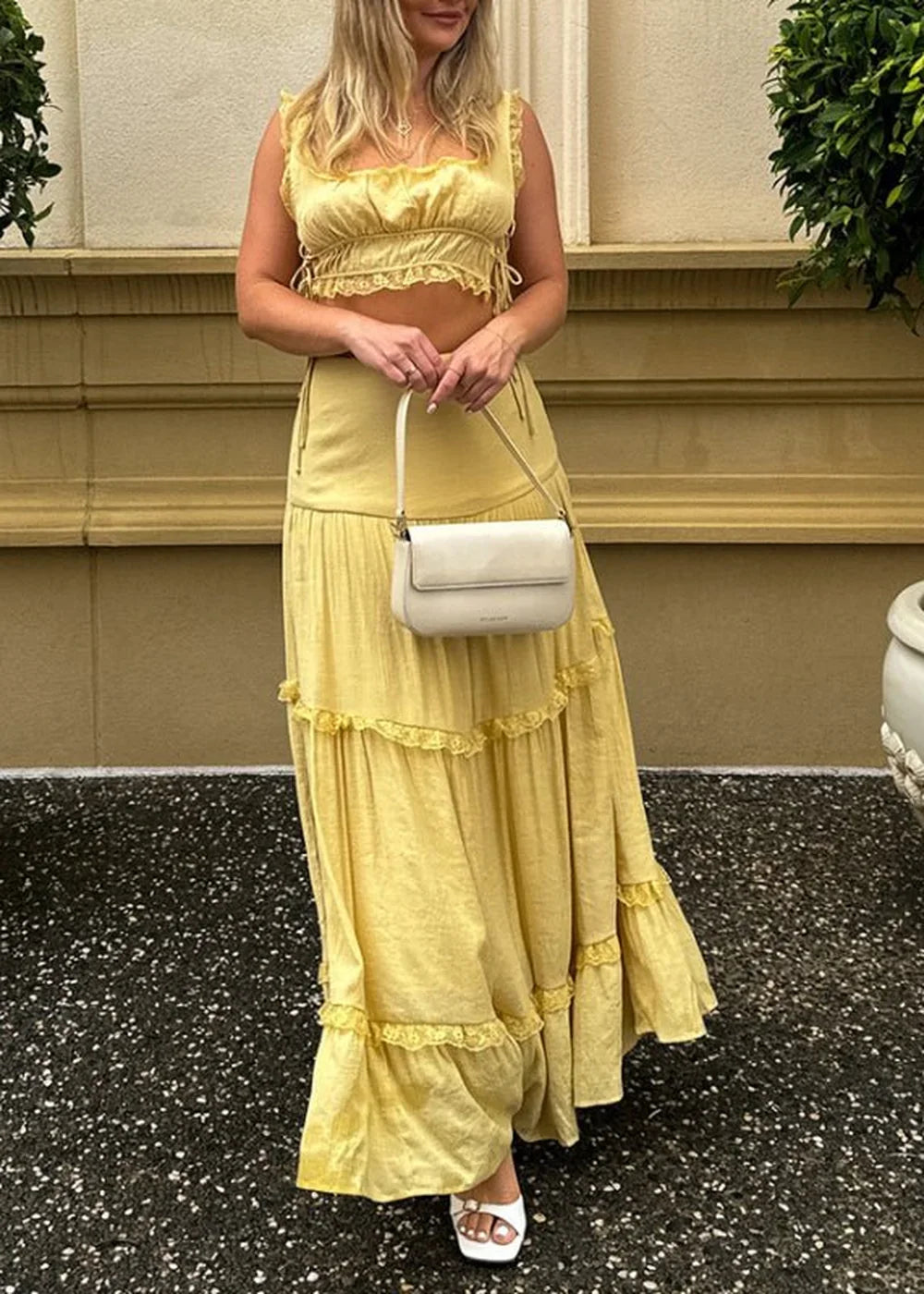 Flowy yellow tiered maxi skirt with lace trim and a matching ruffled crop top, held by a white shoulder bag.