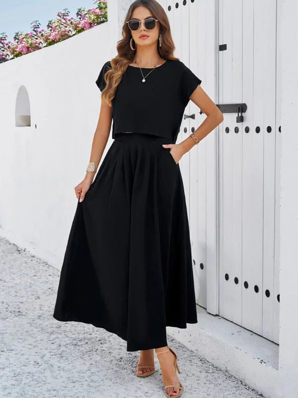 Black two-piece outfit with cropped top and pleated midi skirt on a model, featuring a white wall background.