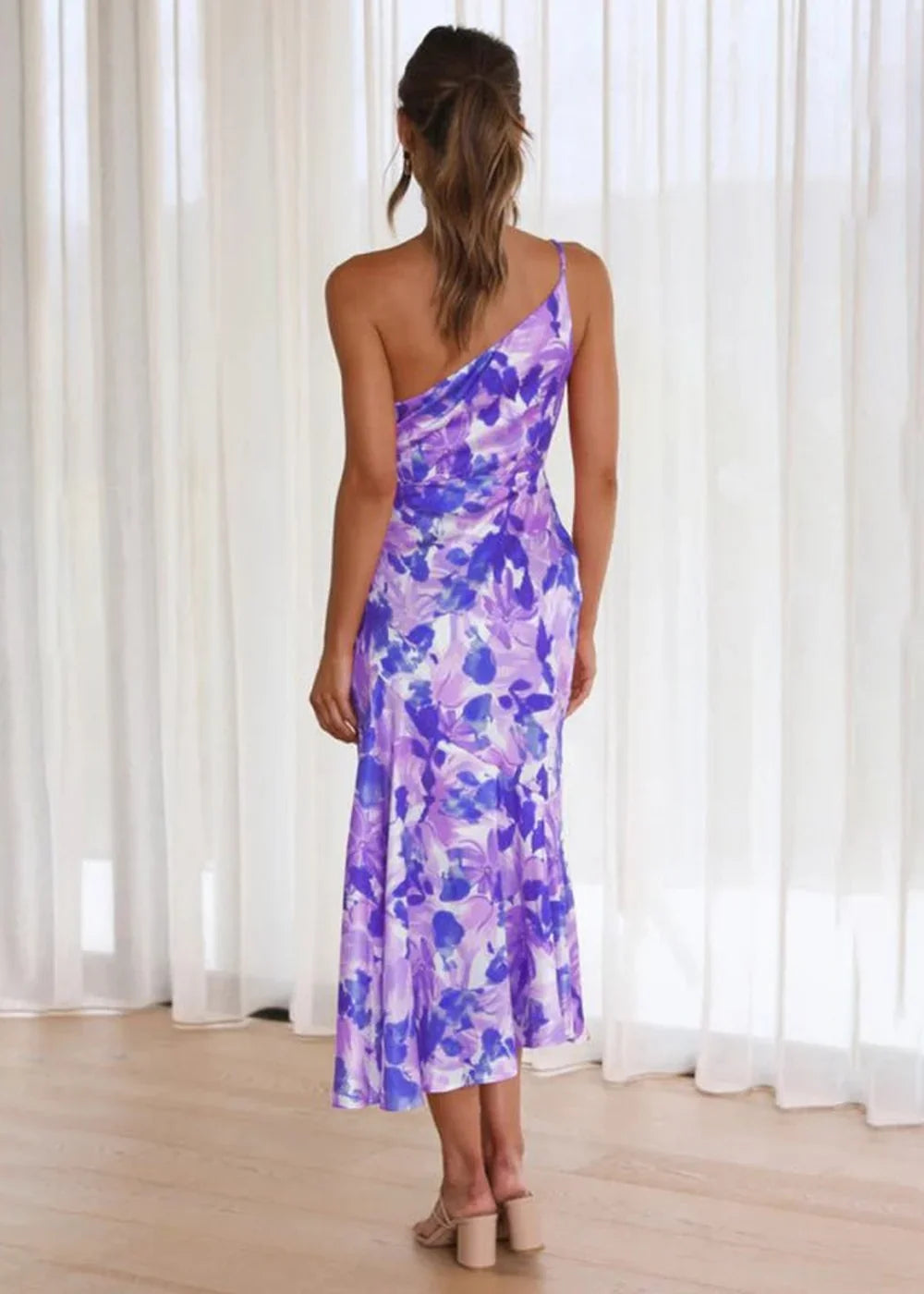 Back view of a woman wearing a purple and white floral print, one-shoulder midi dress.