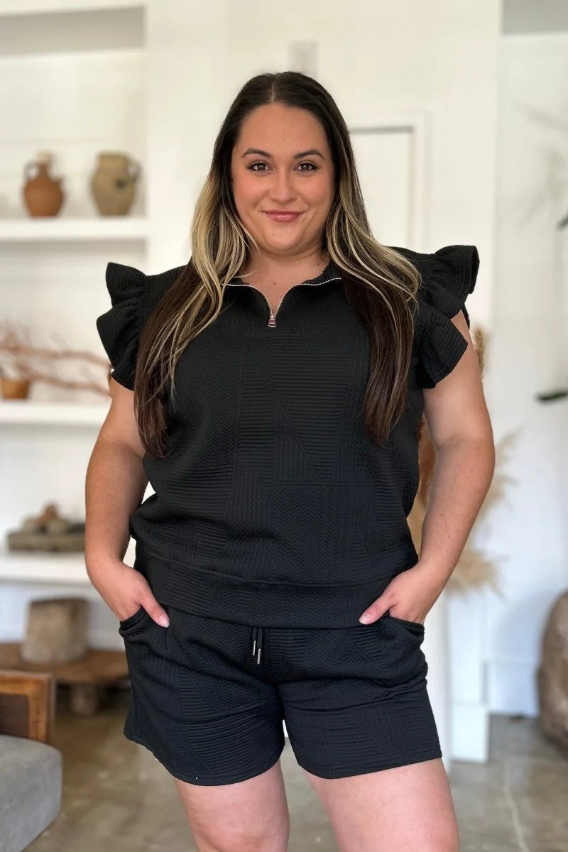 Black textured short suit with ruffled cap sleeves and zip neckline on a woman.