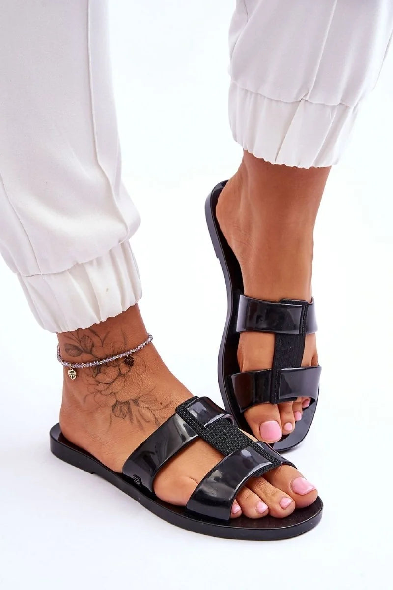 Black plastic slide sandals with double straps on a model's feet, showing a floral tattoo and anklet.