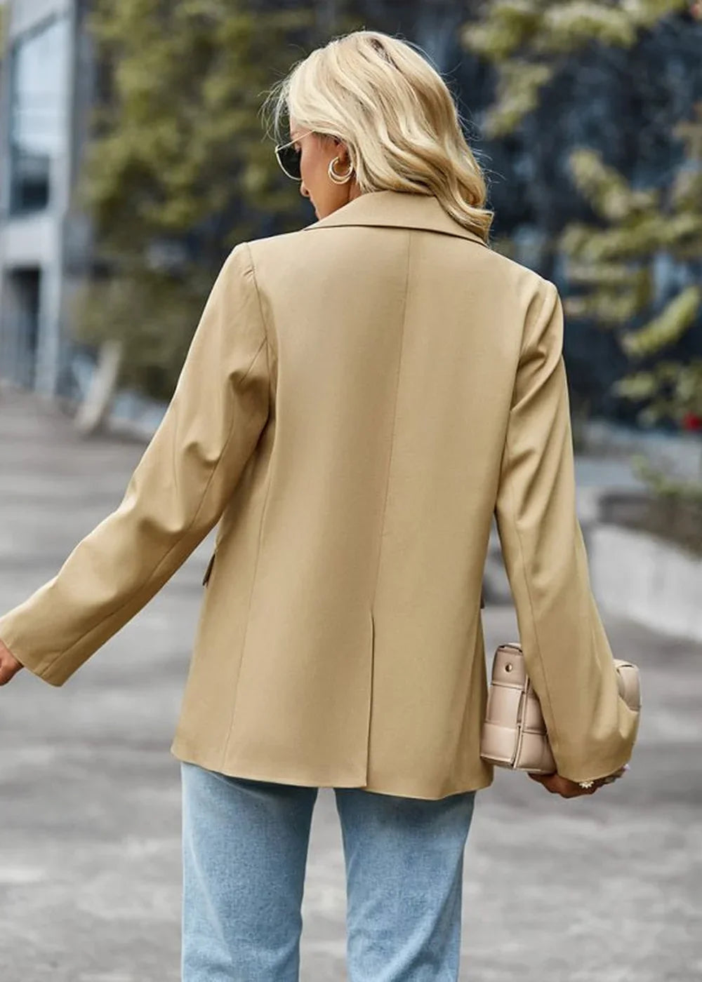 Back view of a woman wearing a tan blazer and light wash jeans, holding a tan handbag.