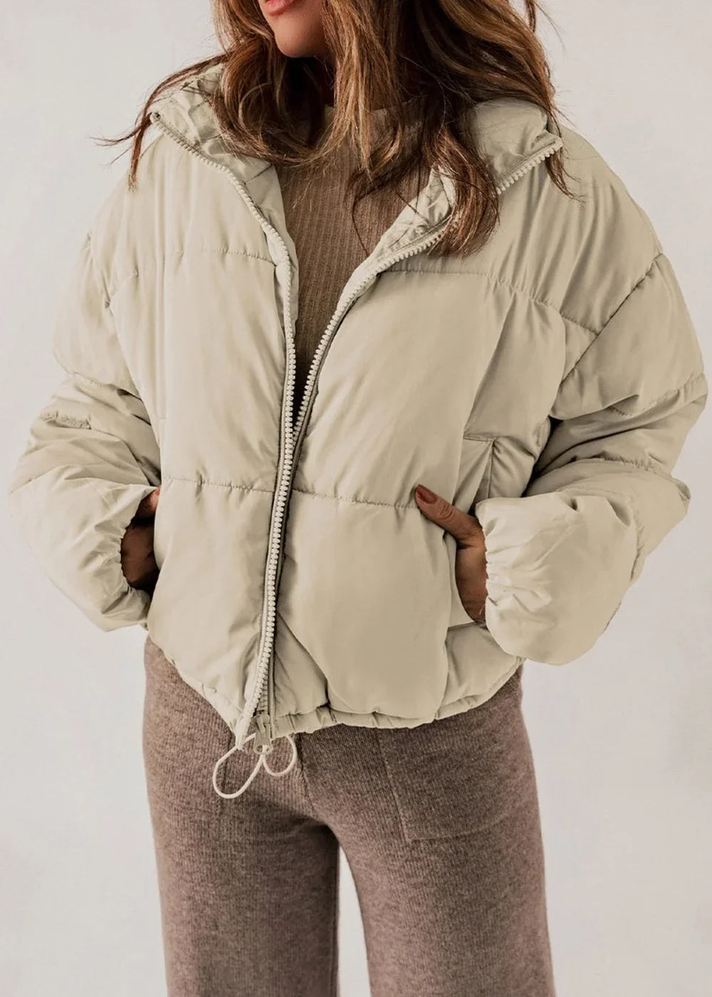 Light beige puffer jacket with hood, zipped front, and pockets, worn over a brown ribbed sweater.
