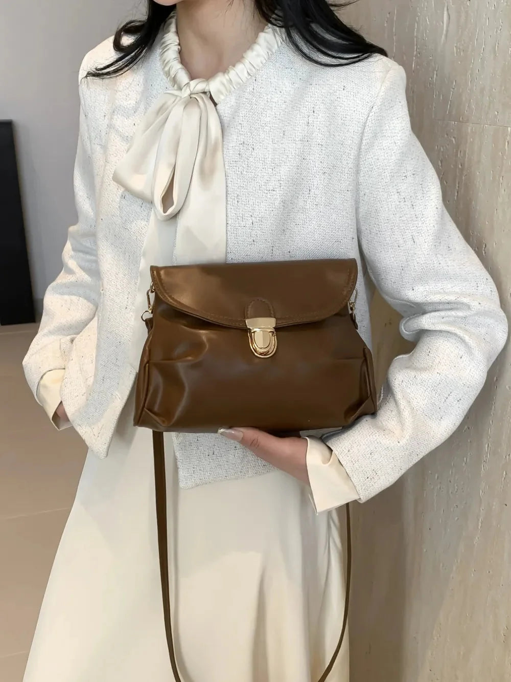 Woman in white tweed jacket and cream dress holds a brown leather crossbody bag with a gold clasp.